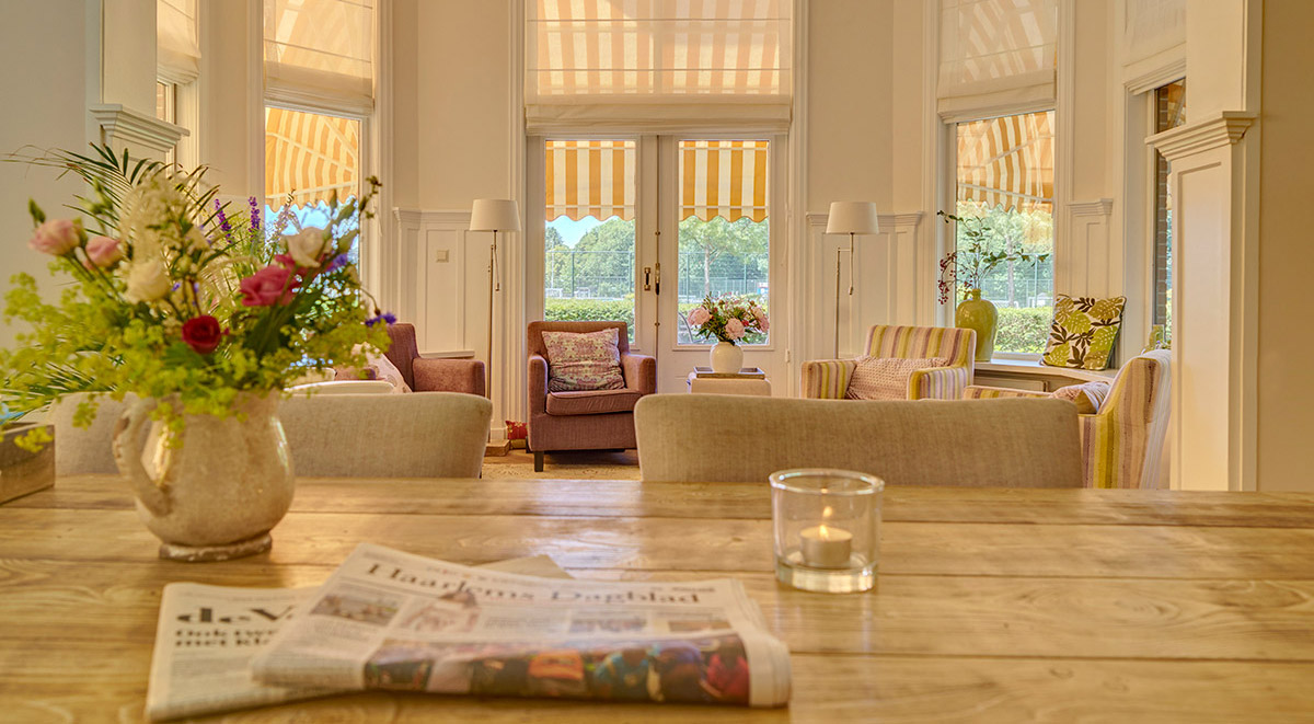 Foto van de woonkamer van het Hospice genomen vanaf de keukentafel met een krant op de voorgrond